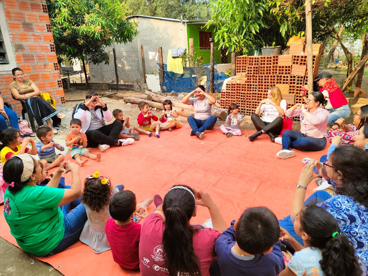 Niñxs y adultos sentados en circulo, poniendo sus manos en corazón frente a su cara.